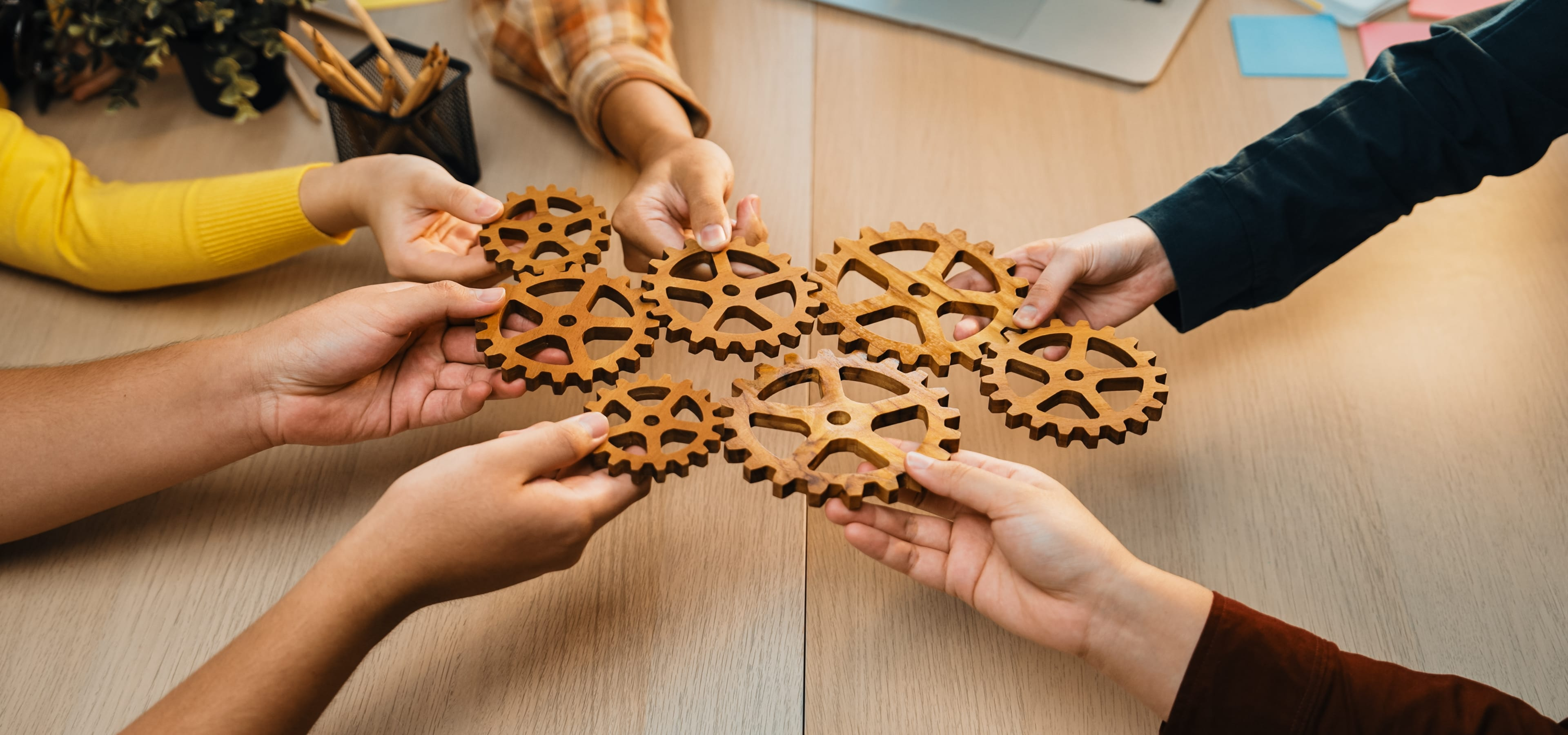 Hands holding interlocked wooden gears on a table, symbolizing teamwork and collaboration.