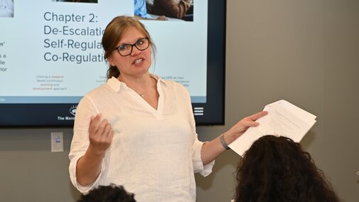 Woman in white shirt giving a presentation to seated audience with slides behind her. "Chapter 2:  De-Escalation and Self-Regulation, Co-Regulation"