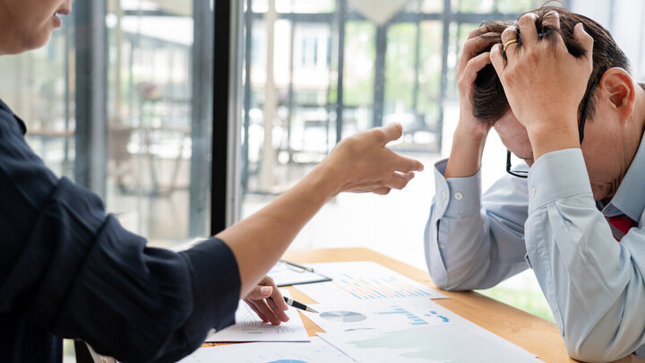 Image of two people sitting on either side of a desk, with one person's head in their hands.