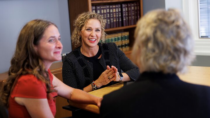 Three women engaged in discussion around a wooden table in an office with bookshelves in the background.