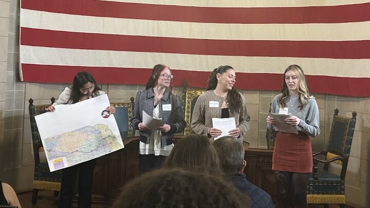 Four young women stand in front of a large American flag, three holding papers and one holding a map, addressing an audience seated before them.
