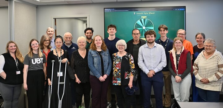 Group of diverse adults posing together in a conference room with a prize wheel displayed on a screen behind them.